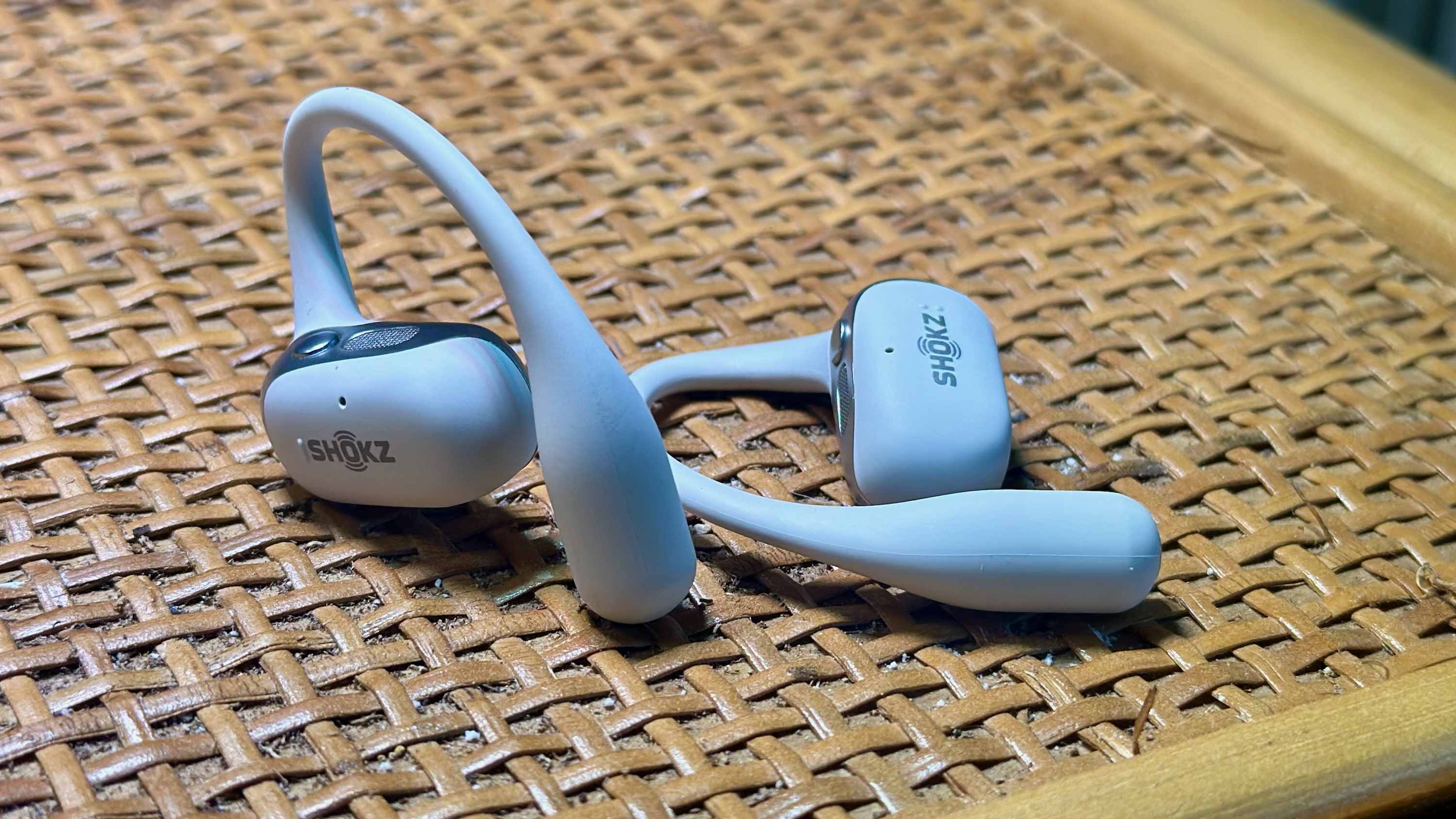 Δύο ακουστικά Shokz OpenFit 2+ σε ξύλινο ράφι, ένα όρθιο και ένα επίπεδο.