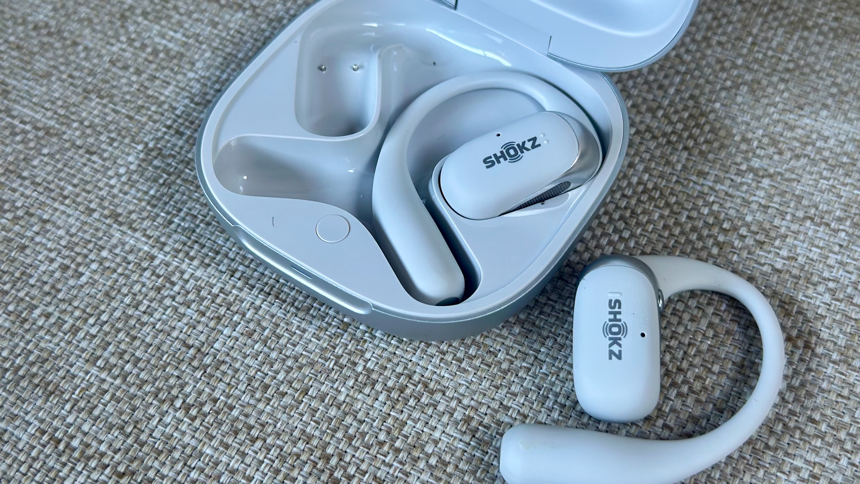 Μια κοντινή φωτογραφία των ακουστικών και της θήκης Shokz OpenFit 2 Plus, το ένα ακουστικό βρίσκεται μέσα και το άλλο δίπλα στη θήκη. Η άδεια υποδοχή φόρτισης και οι μαγνητικές ακίδες είναι ορατές στην αριστερή πλευρά της θήκης.