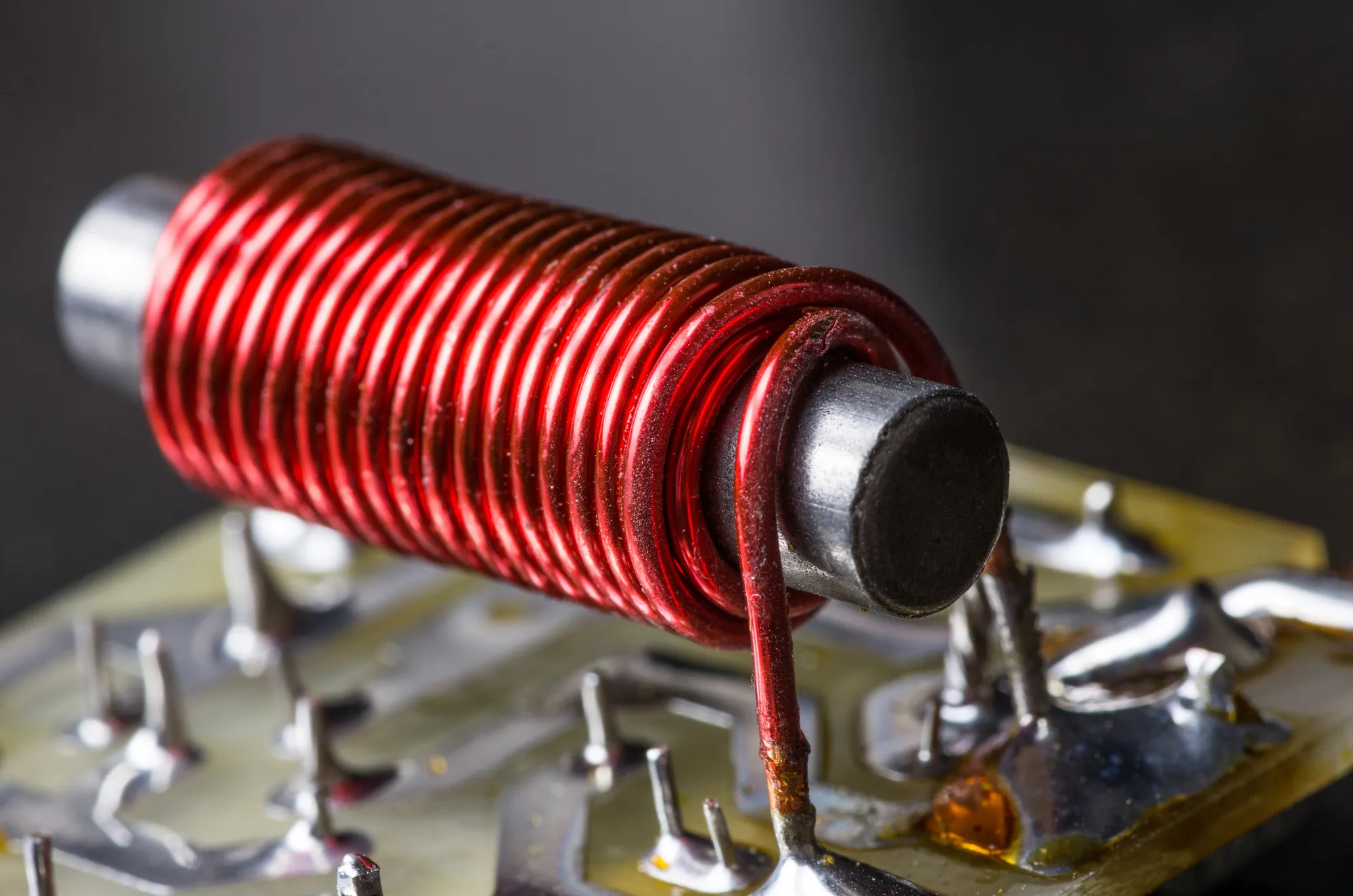 
A close-up photograph of an electronic component, which appears to be an electromagnet or inductor. It consists of a shiny, metallic cylindrical core wrapped tightly with red insulated copper wire, and is mounted on a circuit board.