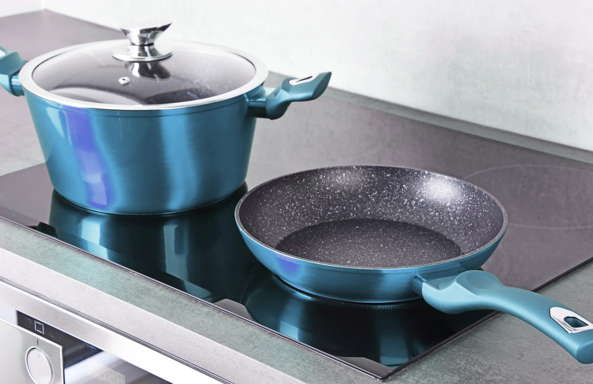 
A close-up shot of a modern, black induction cooktop with two pieces of cookware: a large, teal-blue stockpot with a glass lid, and a matching teal-blue frying pan with a dark, non-stick interior. The glossy surface of the cooktop reflects the pots.