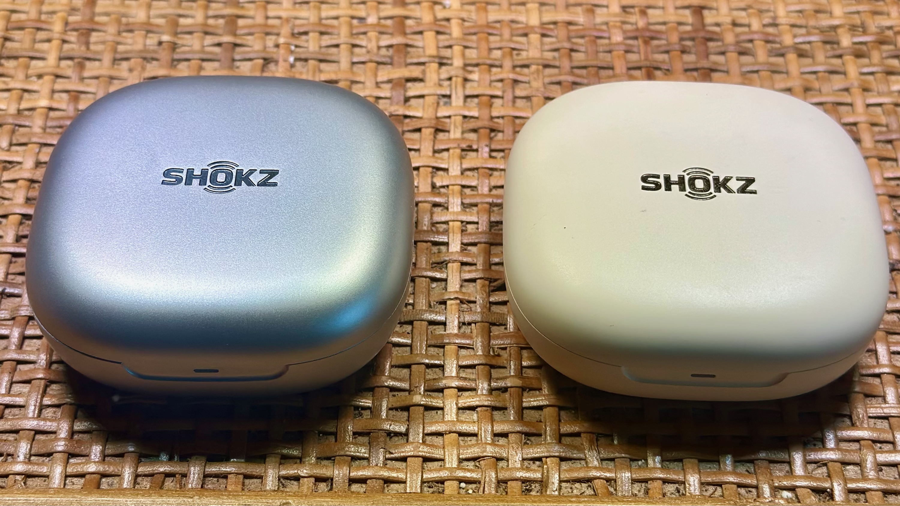 Μια κλειστή ασημί θήκη Shokz OpenFit 2+ (αριστερά) δίπλα σε μια κλειστή λευκή θήκη Shokz OpenFit 2 (δεξιά).
