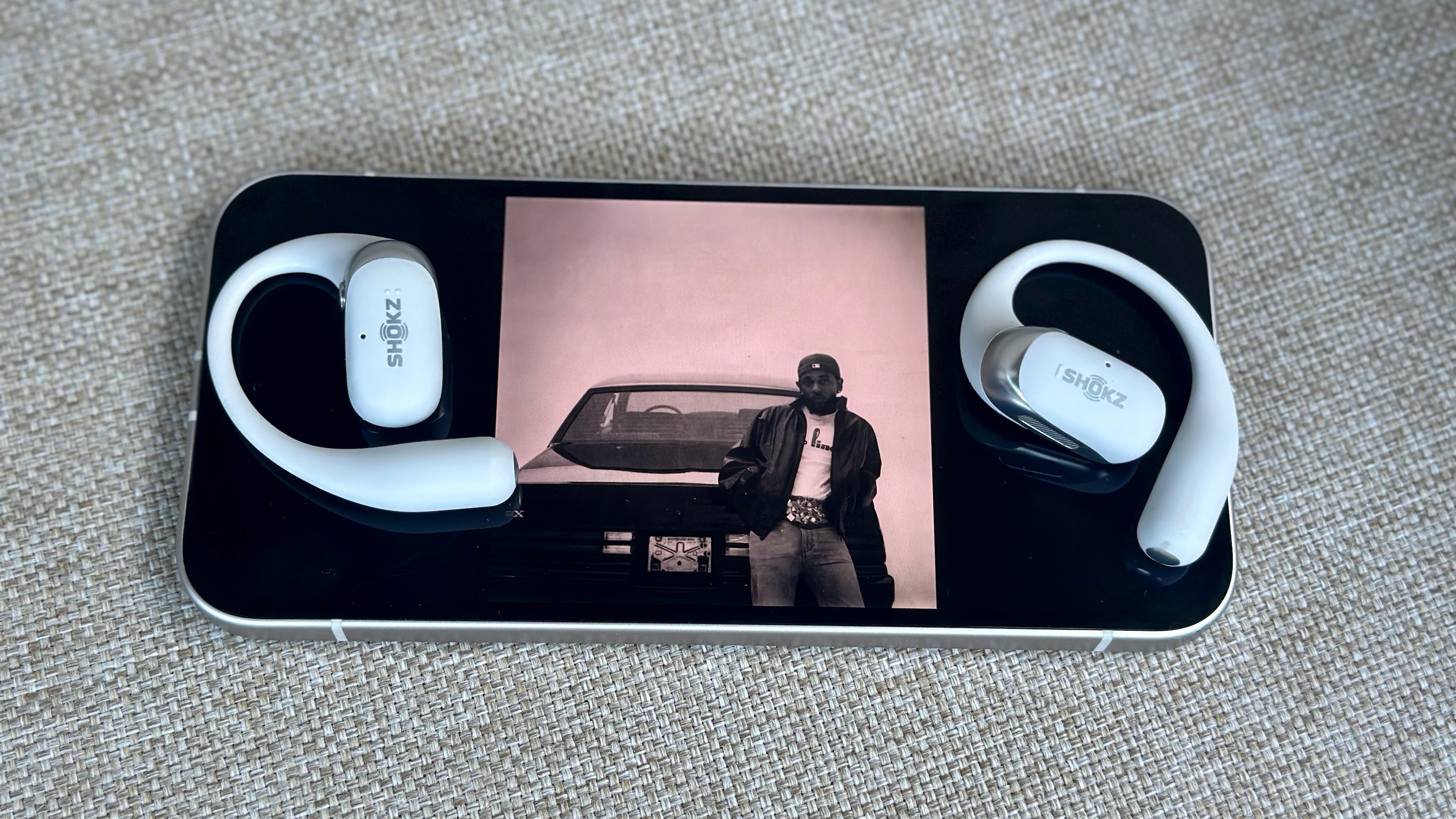 Δύο ακουστικά Shokz OpenFit 2 Plus που βρίσκονται πάνω από ένα Google Pixel 9a, το οποίο εμφανίζει ένα στιγμιότυπο οθόνης ενός άλμπουμ Kendrick Lamar που παίζει πάνω από τα ακουστικά.