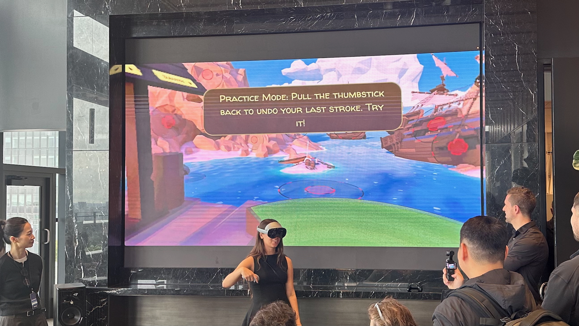 Μια φωτογραφία ενός μηχανικού της Samsung που παίζει Walkabout Mini Golf ενώ φοράει τα ακουστικά Galaxy XR και χρησιμοποιεί ένα χειριστήριο Galaxy XR στο δεξί της χέρι, μια οθόνη που δείχνει το παιχνίδι πίσω της.