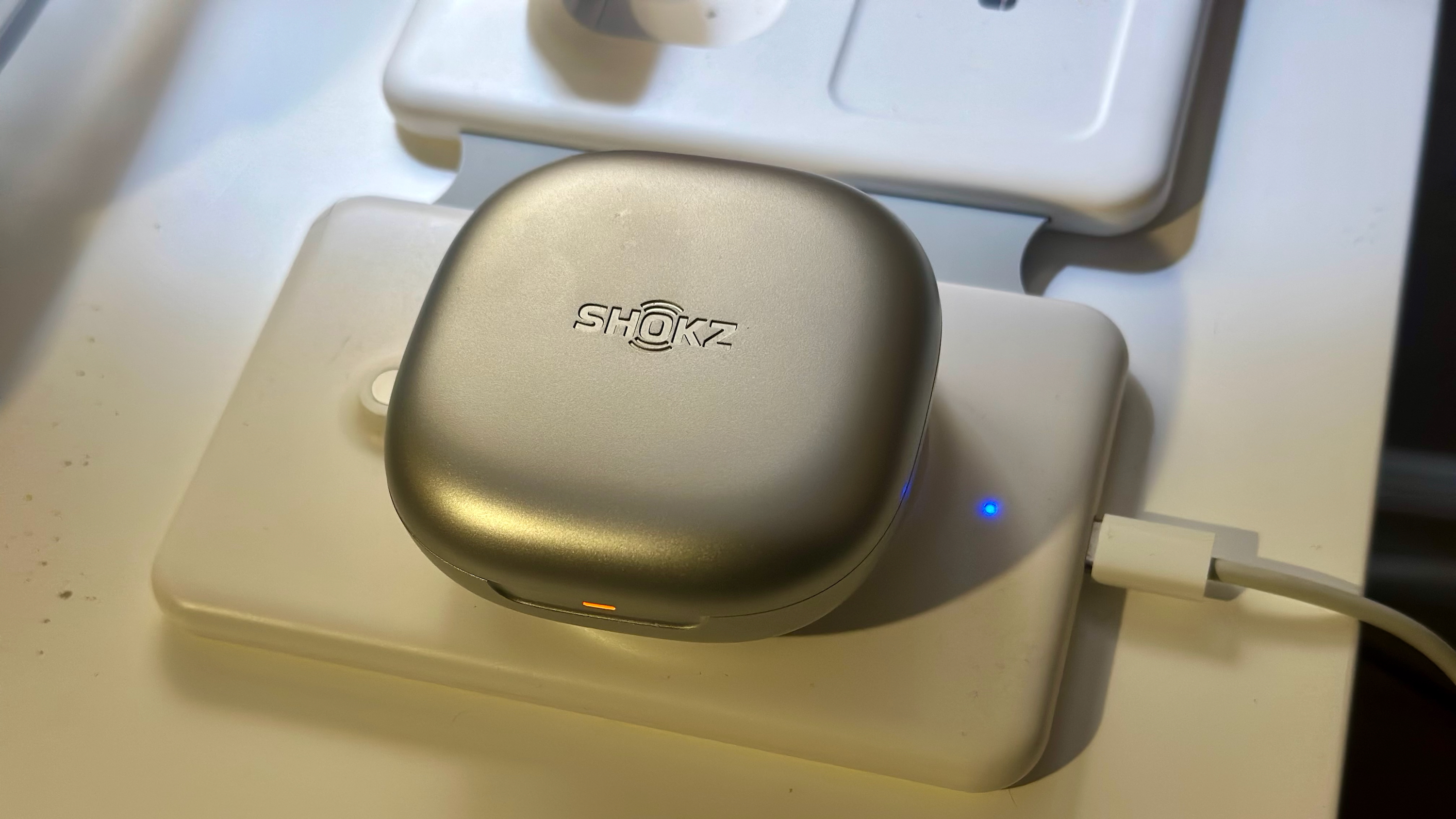 Μια φωτογραφία της κλειστής θήκης Shokz OpenFit 2 Plus που βρίσκεται πάνω από έναν φορτιστή Qi, με ορατή την πορτοκαλί ένδειξη φόρτισης.