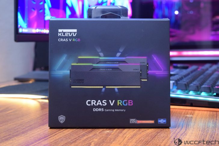 The image shows a box of KLEVV CRAS V RGB DDR5 Gaming Memory placed on a wooden desk, with a colorful background and a keyboard partially visible.