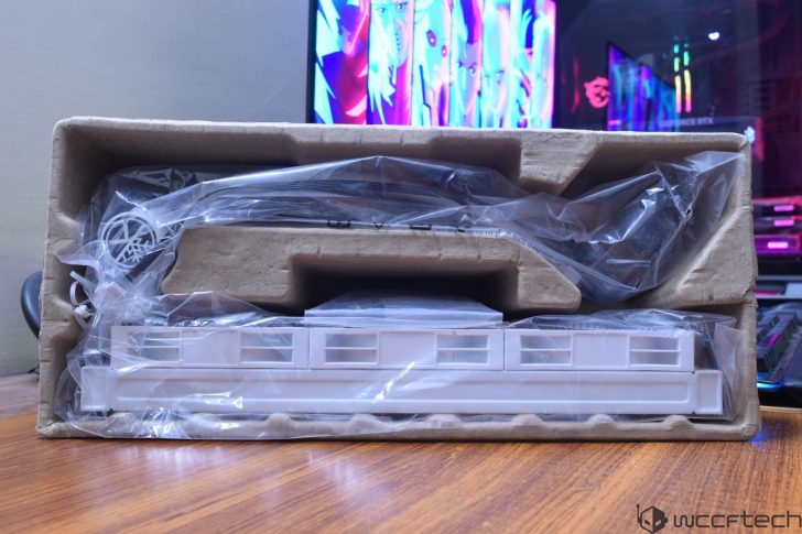 Μια λευκή κάρτα γραφικών χωρίς επωνυμία είναι τυλιγμένη σε πλαστικό μέσα σε ένα δίσκο συσκευασίας από χαρτόνι, με επιφάνεια εργασίας παιχνιδιών με φωτισμό RGB και ζωντανή οθόνη στο φόντο.