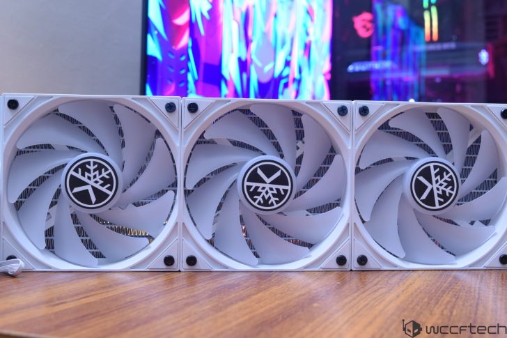 Μια κινηματογράφηση σε πρώτο πλάνο τριών ανεμιστήρων ψύξης υπολογιστή με επωνυμία Snowflake, τακτοποιημένα δίπλα-δίπλα σε μια ξύλινη επιφάνεια, με μια πολύχρωμη οθόνη οθόνης στο φόντο.