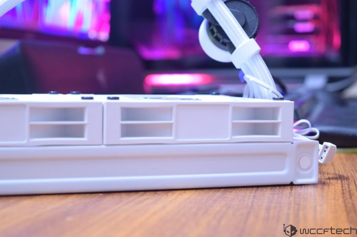 Ένα κοντινό πλάνο ενός λευκού καλοριφέρ υπολογιστή με προσαρτημένους σωλήνες, τοποθετημένο σε ξύλινη επιφάνεια με πολύχρωμα φώτα LED στο φόντο.