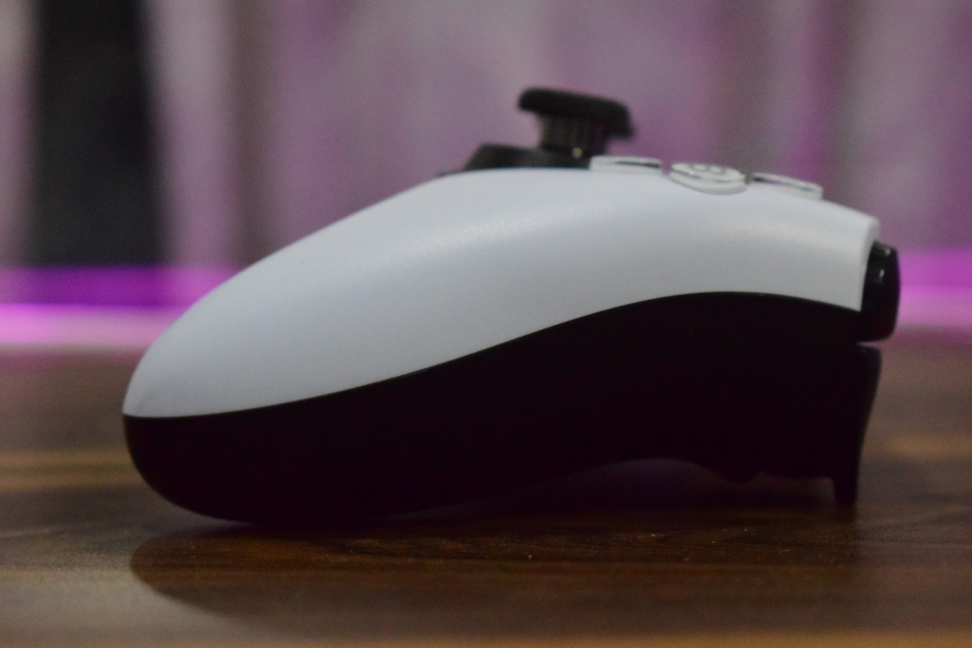 Ικανοποιητικός ελεγκτής προϋπολογισμού για PC και Xbox Λευκό και μαύρο χειριστήριο PlayStation 5 DualSense πλάγια όψη σε ξύλινη επιφάνεια με θολό ροζ φόντο.