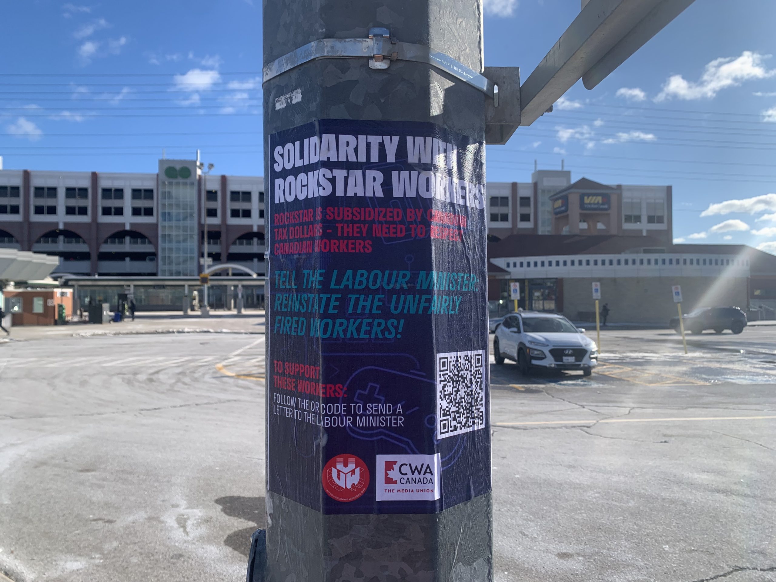 CWA Canada and Local Toronto Game Devs Protest at Rockstar Toronto in Solidarity with 34 Fired Workers A poster on a pole in a parking lot reads 'Solidarity with Rockstar Workers,' urging the public to pressure the labor minister to reinstate fired workers, with a QR code and CWA Canada logo visible.