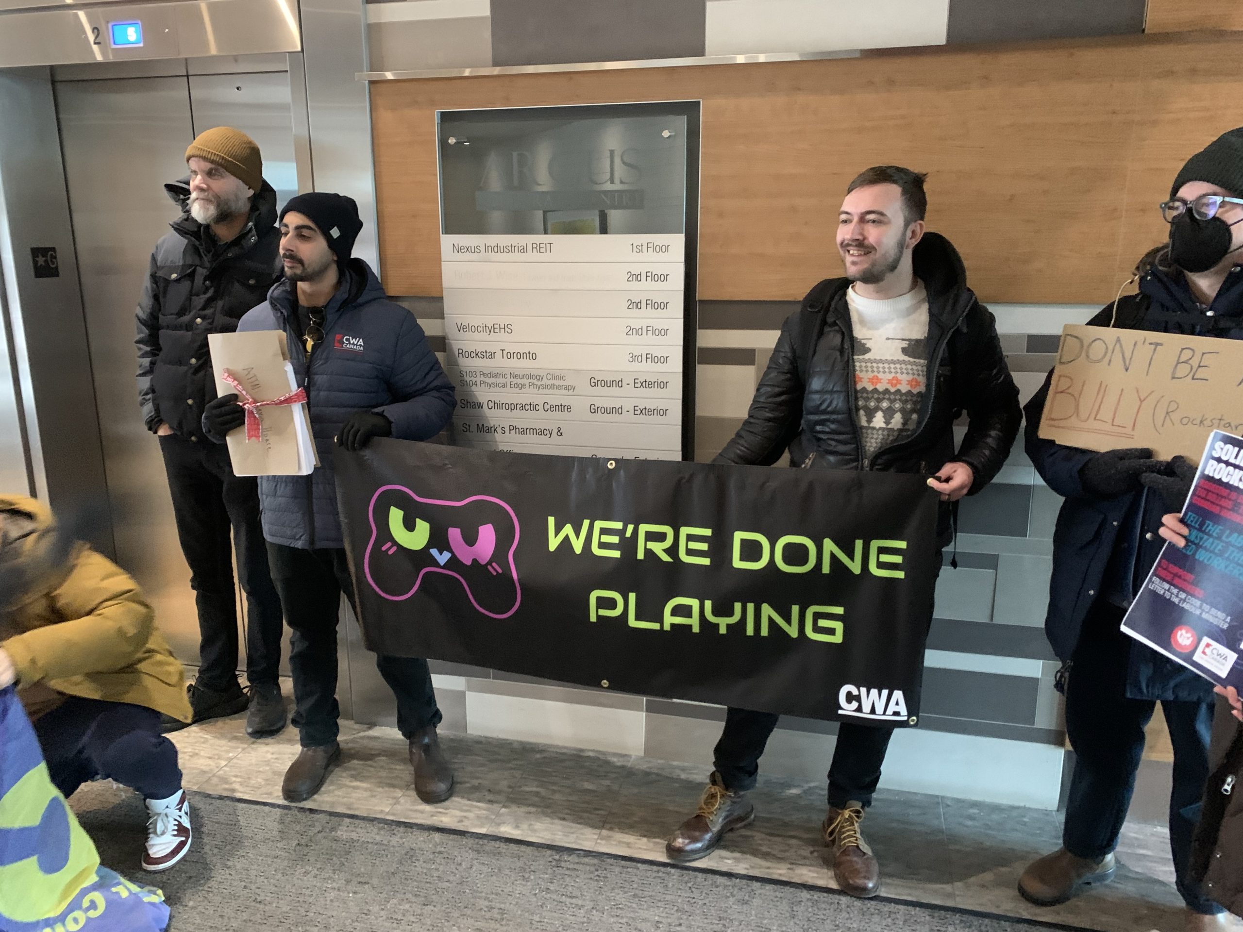 CWA Canada and Local Toronto Game Devs Protest at Rockstar Toronto in Solidarity with 34 Fired Workers People are standing in front of an office directory sign at Nexus Industrial REIT, holding a banner with a game controller icon that reads 'WE'RE DONE PLAYING' and signs mentioning 'Rockstar Toronto' and 'DON'T BE A BULLY'.