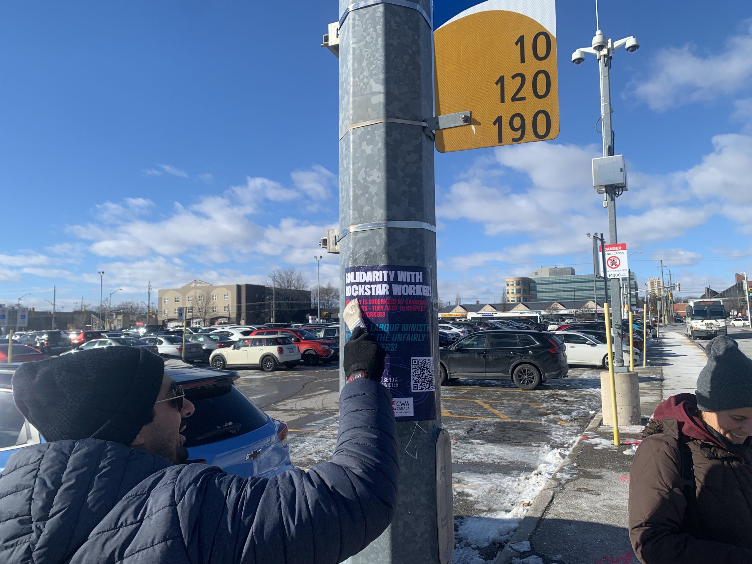 CWA Canada and Local Toronto Game Devs Protest at Rockstar Toronto in Solidarity with 34 Fired Workers A person wearing a winter jacket and beanie is removing a poster titled 'Solidarity with Rockstar Workers' from a snow-dusted parking lot signpost.