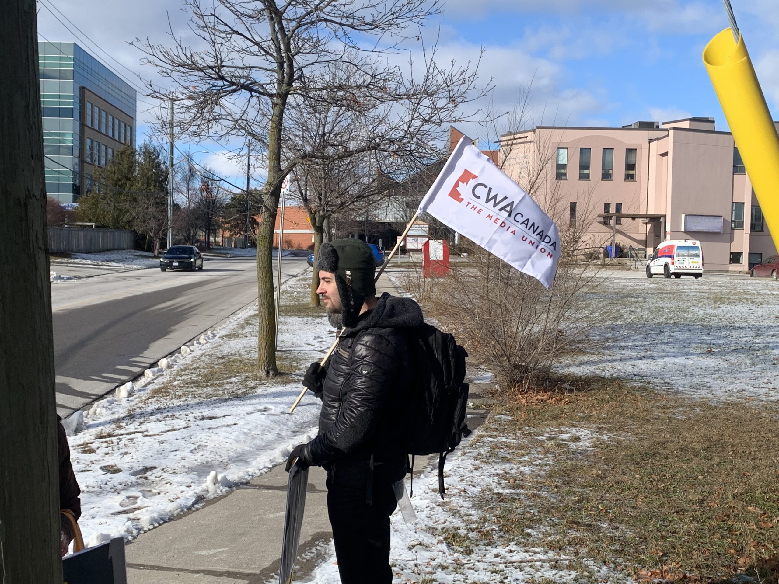 CWA Canada and Local Toronto Game Devs Protest at Rockstar Toronto in Solidarity with 34 Fired Workers A person stands outside holding a CWA Canada 'THE MEDIA UNION' flag near a snowy street with a building and parked van in the background.