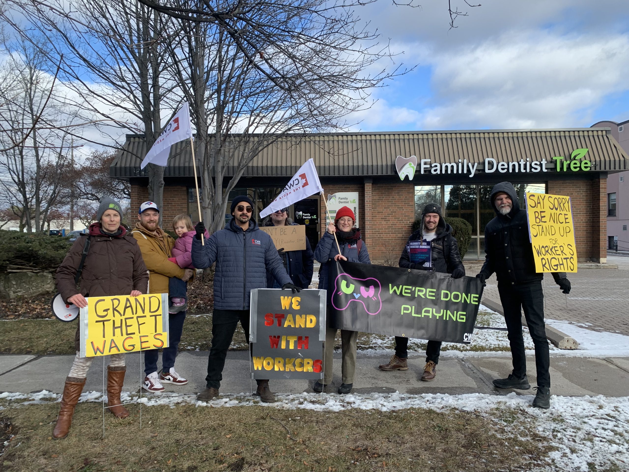 CWA Canada and Local Toronto Game Devs Protest at Rockstar Toronto in Solidarity with 34 Fired Workers A group of people standing outside a building labeled Family Dentist Tree hold signs saying 'Grand Theft Wages,' 'We Stand With Workers,' and 'Say Sorry Be Nice Stand Up For Worker's Rights,' with some carrying flags bearing the logo CWA.