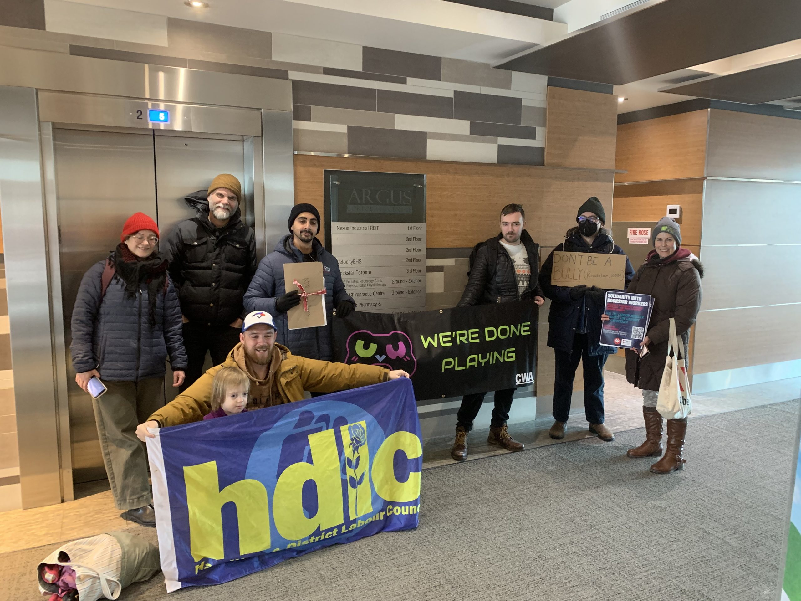 CWA Canada and Local Toronto Game Devs Protest at Rockstar Toronto in Solidarity with 34 Fired Workers A group of people inside a building holding signs that read 'HDC,' 'WE'RE DONE PLAYING,' and 'DON'T BE A BULLY,' with some individuals smiling and others holding informational flyers.