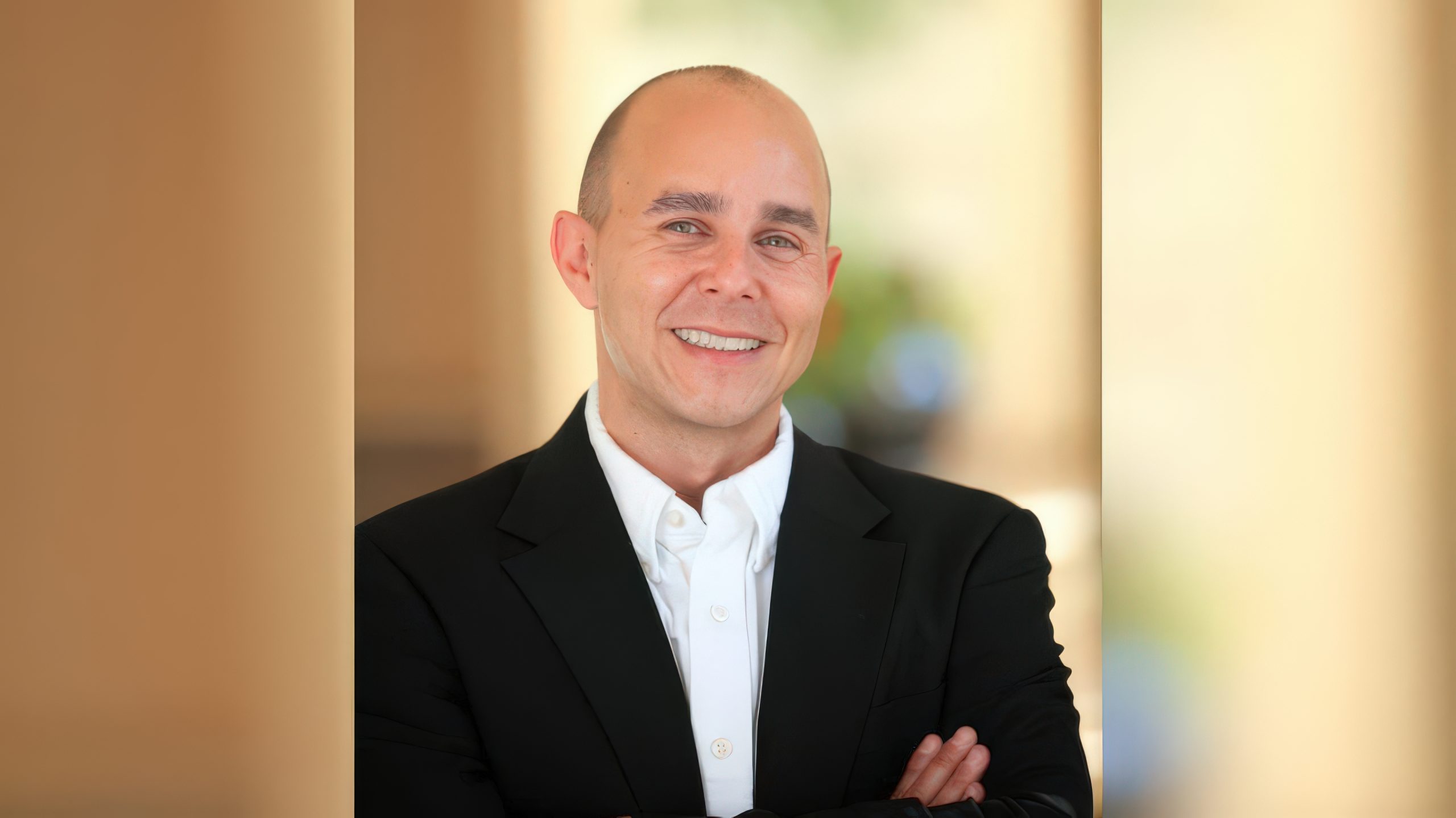 Is the MMO Genre Dead for Western AAA Developers? We Asked Three Industry Veterans A person in a black suit with a white shirt smiling in a blurred indoor setting.