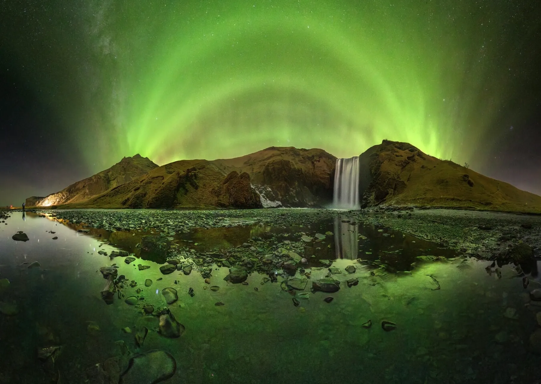 a bright green aurora over a waterfall and reflecting in a stony pool