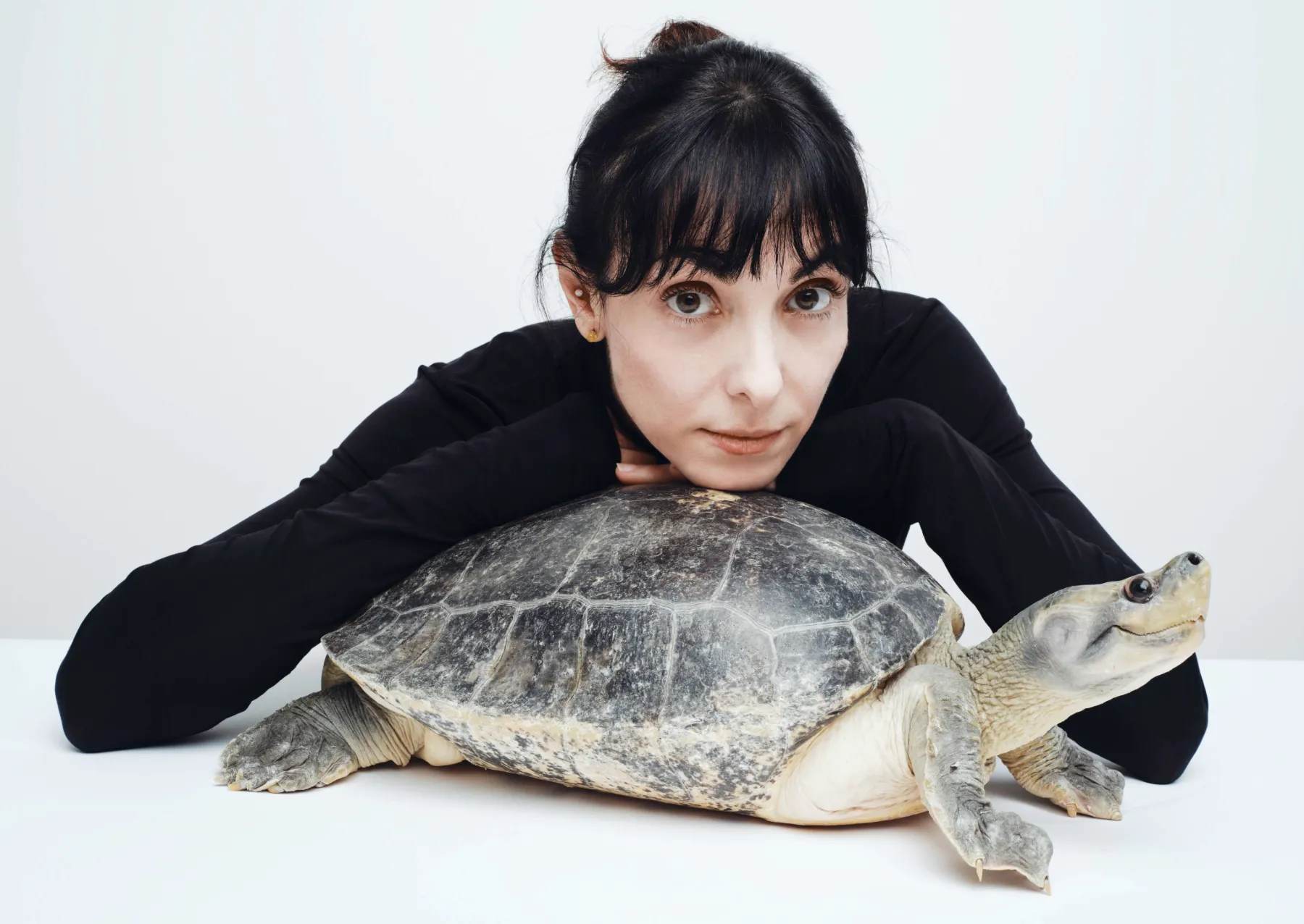μια γυναίκα με μαύρο λαιμό χελώνας ποζάρει με μια μεγάλη χελώνα με γυρισμένη μύτη