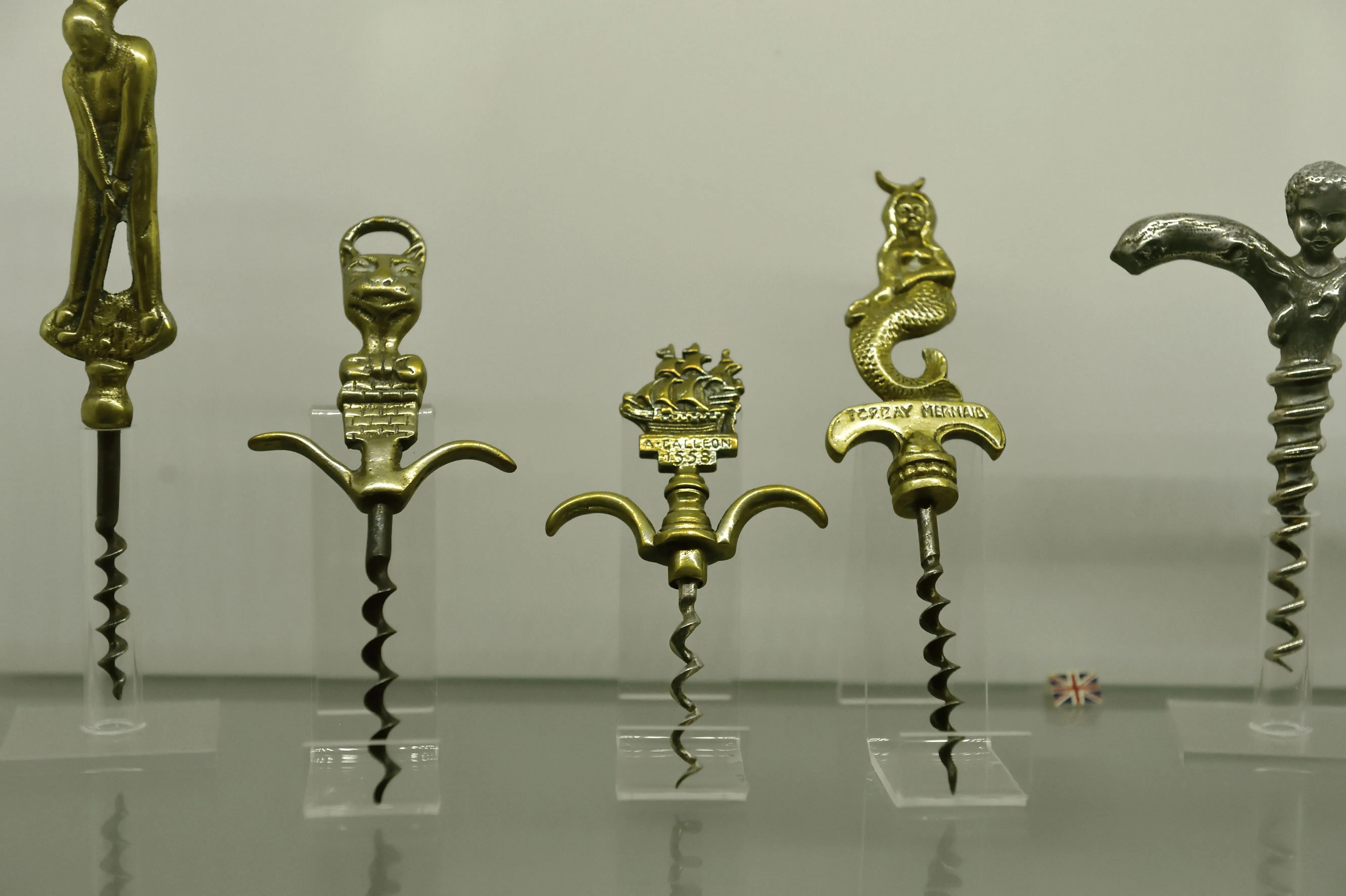 Μια σειρά από πέντε περίτεχνα vintage τιρμπουσόν που εμφανίζονται σε διαφανές ακρυλικό στέκεται σε ένα απλό λευκό φόντο. Οι λαβές είναι κατασκευασμένες από ορείχαλκο και ασήμι σε διάφορες φιγούρες, όπως: Ένας παίκτης γκολφ στη μέση.  Ένα στυλιζαρισμένο κεφάλι γάτας ή γαργκόιλ.  Ένα πλοίο γαλέρας με ετικέτα "A. Galleon 1588."Μια γοργόνα με ετικέτα "Torquay Mermaid."Μια φιγούρα χερουβείμ που σχηματίζει τη λαβή μιας σπείρας σε ασημί τόνους.
