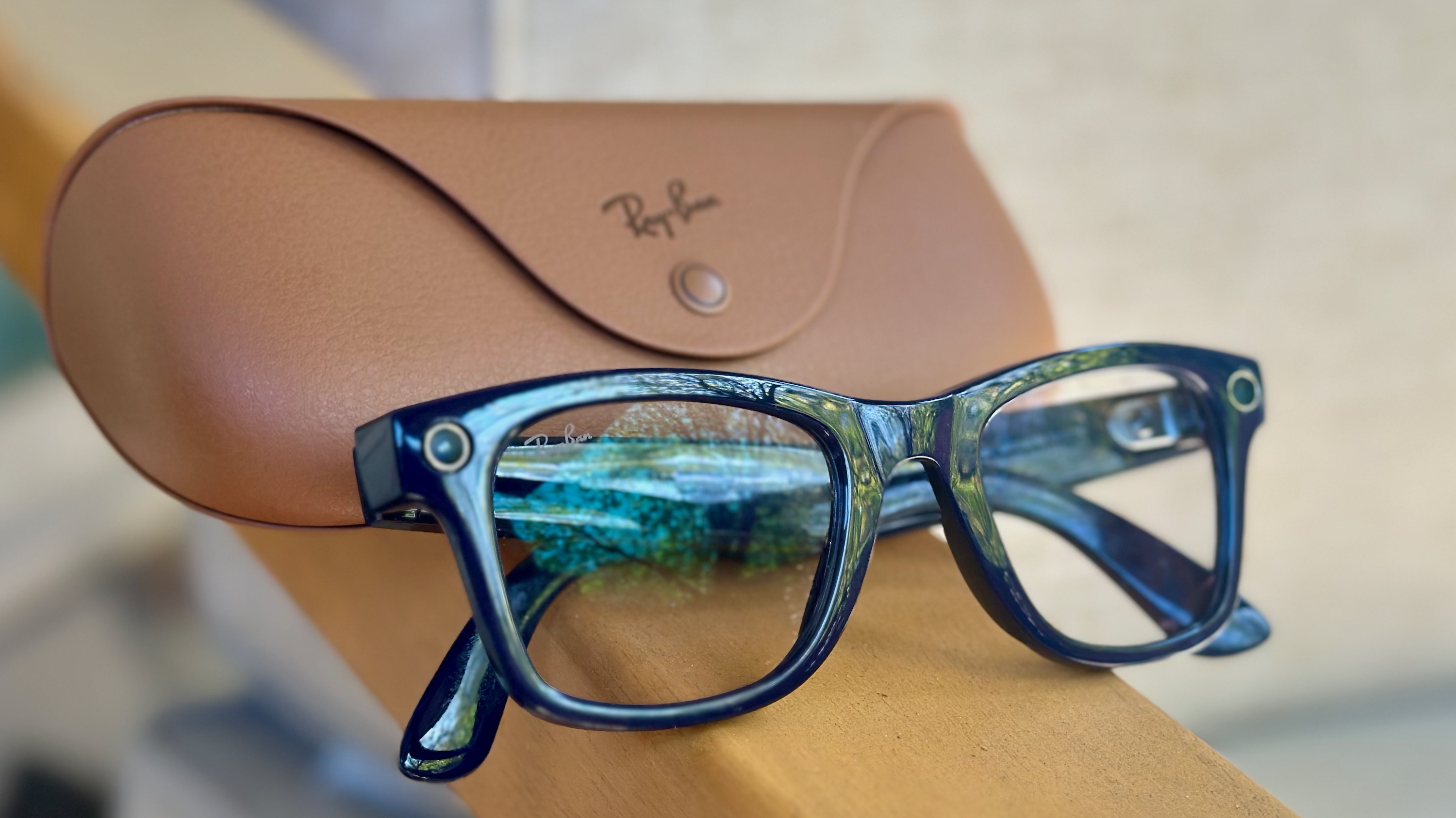 Κοντινή φωτογραφία των γυαλιών Wayfarer Shiny Cosmic Blue Ray-Ban Meta (Gen 2) διπλωμένα σε ένα υποβραχιόνιο με την επίσημη καφέ θήκη φόρτισης Ray-Ban πίσω του.