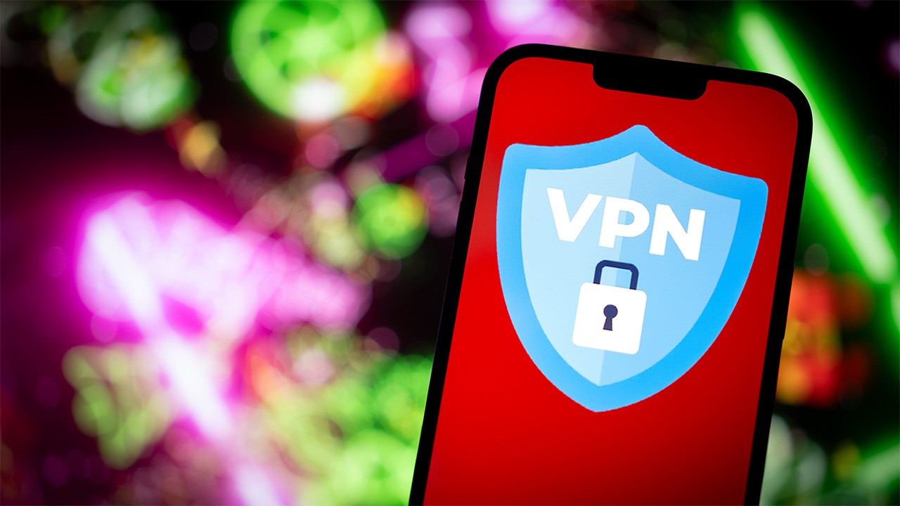Smartphone που εμφανίζει μπλε λογότυπο ασπίδας VPN με λουκέτο. Υπάρχει ένα πράσινο και ροζ θολό τεχνολογικό φόντο