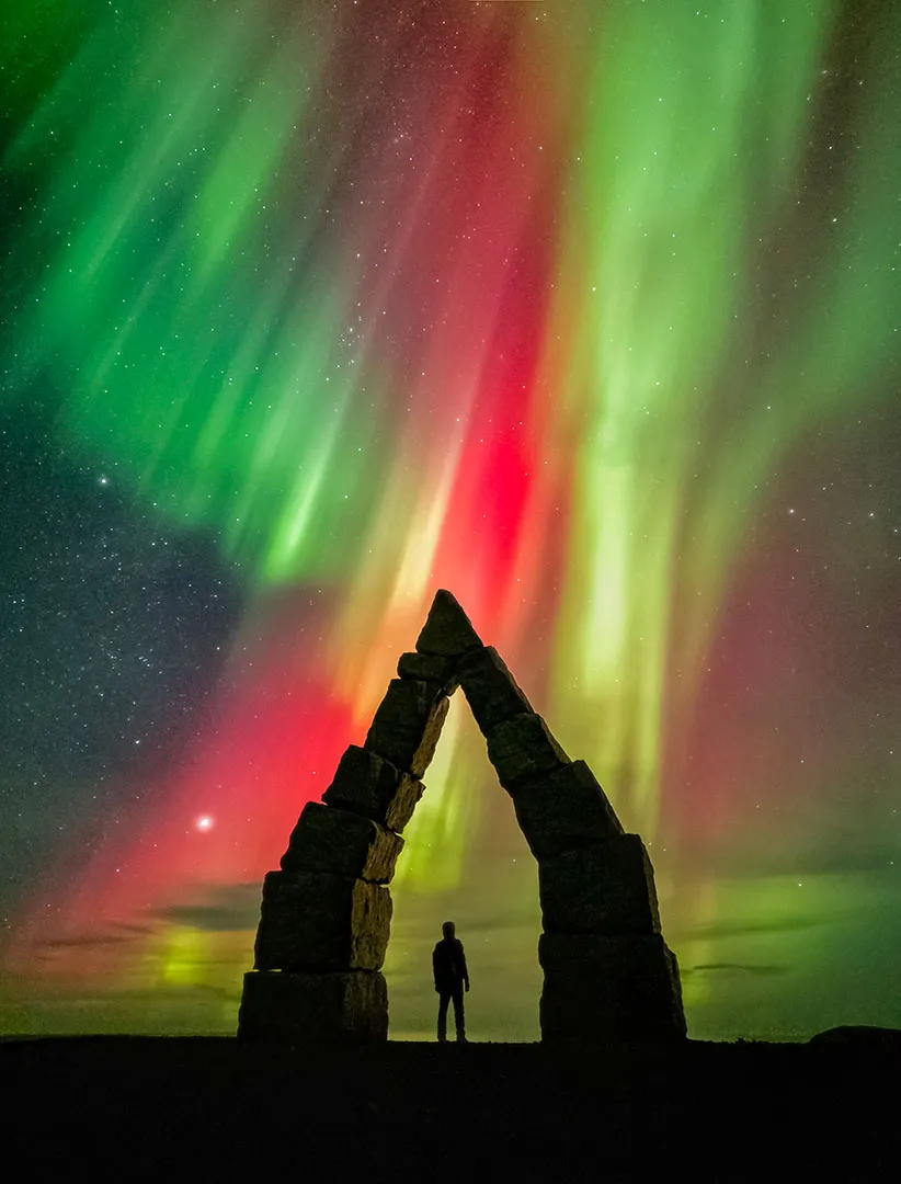 a man stands under an arch while a red and green aurora dances in the night sky