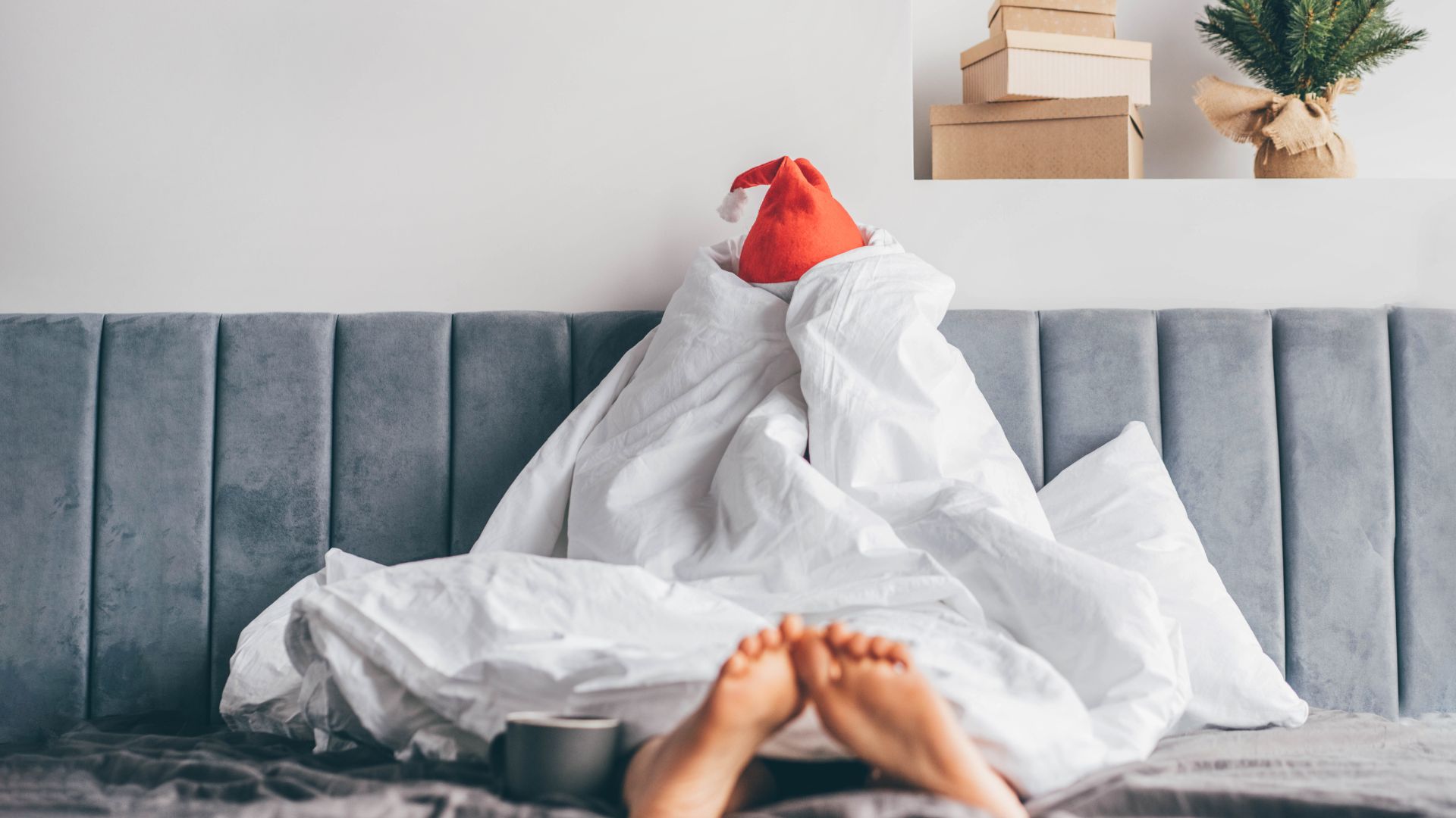 Ένα άτομο που κάθεται στο κρεβάτι τυλιγμένο με ένα λευκό πάπλωμα που καλύπτει ολόκληρο το σώμα του εκτός από τα πόδια του. Ένα καπέλο Άγιου Βασίλη φαίνεται να βγαίνει από το πάνω μέρος του παπλώματος.