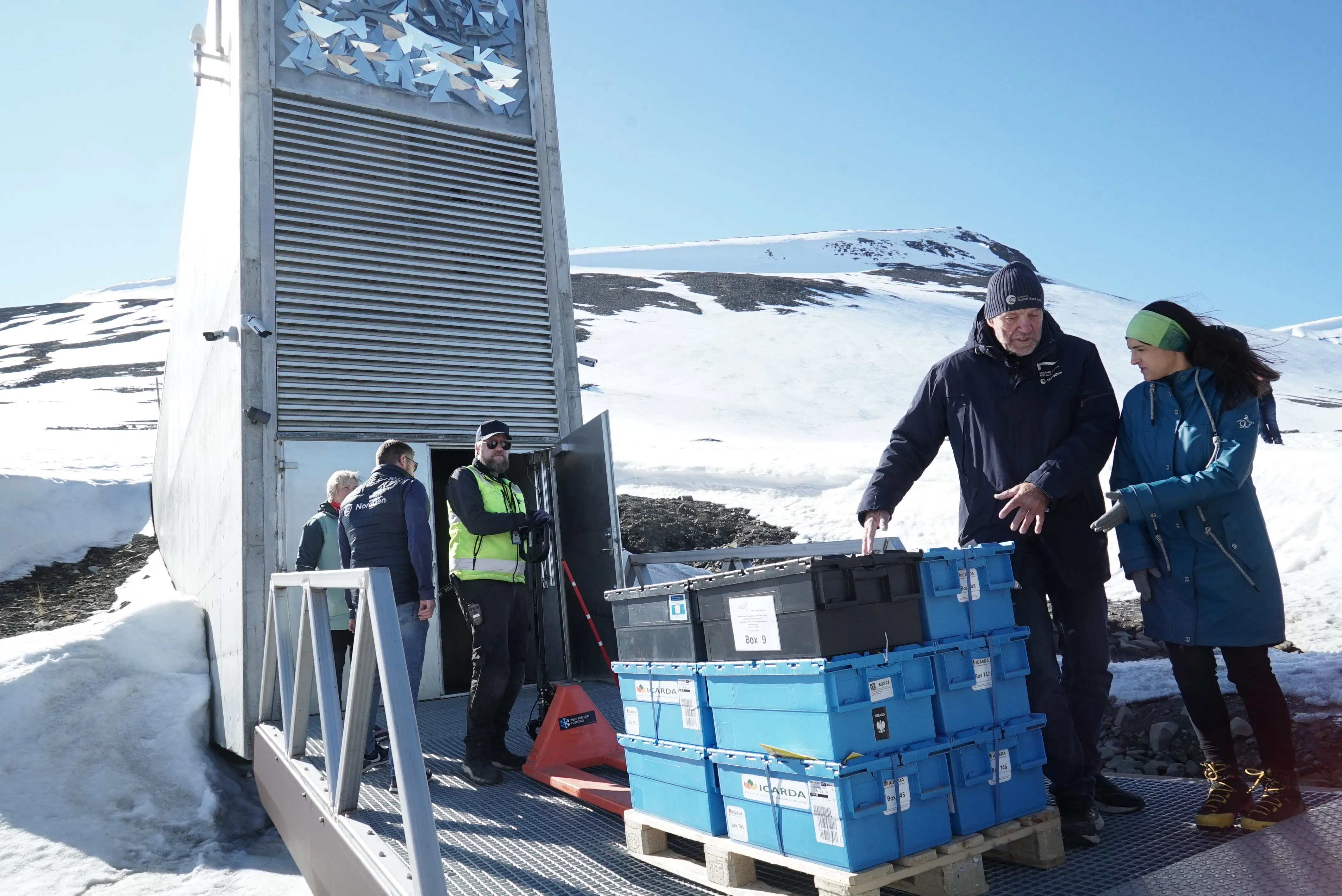 Η τσιμεντένια είσοδος του Svalbard Global Seed Vault βρίσκεται φωλιασμένη σε μια χιονισμένη βουνοπλαγιά κάτω από έναν καταγάλανο ουρανό. Σε πρώτο πλάνο, πολλά άτομα είναι συγκεντρωμένα σε ένα μεταλλικό διάδρομο κοντά στην ανοιχτή πόρτα του θησαυρού. Μια στοίβα από μπλε και μαύρα κιβώτια αποθήκευσης, με ετικέτα το "ICARDA" λογότυπο, στηρίζεται σε μια ξύλινη παλέτα καθώς ένας άνδρας σε σκοτεινό πάρκο τους κάνει χειρονομίες.