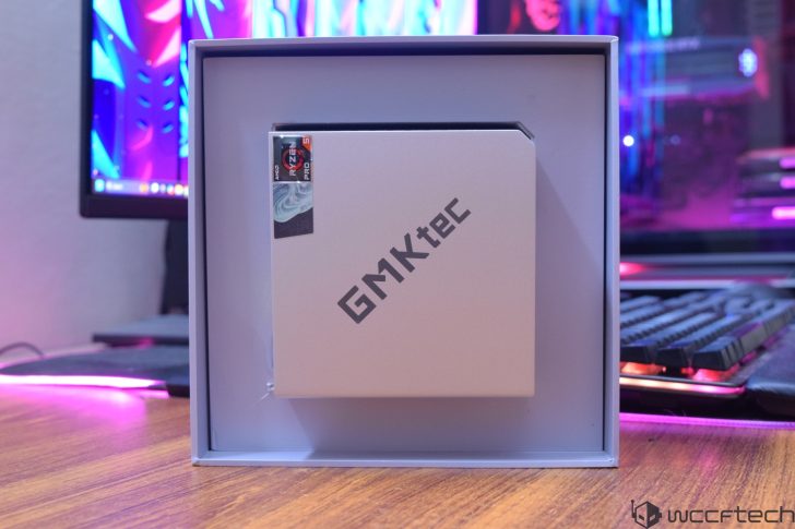 A GMKtec box with an AMD Ryzen 5 Pro sticker is displayed on a desk in front of a gaming setup with a colorful monitor and illuminated keyboard.