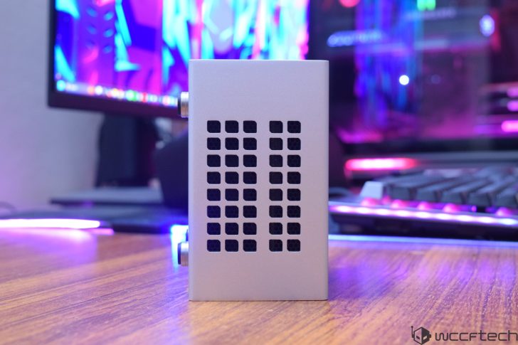 A small, unbranded silver enclosure with ventilation holes is displayed on a wooden desk, with a brightly lit computer setup in the background.