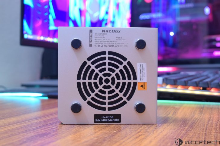 The bottom view of a GMKtec NucBox mini PC model M6 is shown on a wooden desk with a colorful monitor in the background.