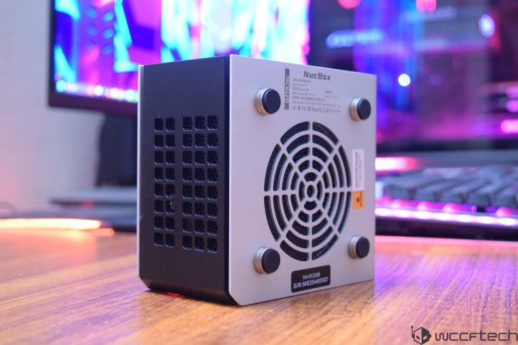 A GMKtec NucBox mini PC is positioned on a wooden desk in front of a blurred monitor and keyboard, with visible text on the front reading '16+512GB' and 'S/N: MB252440387'.