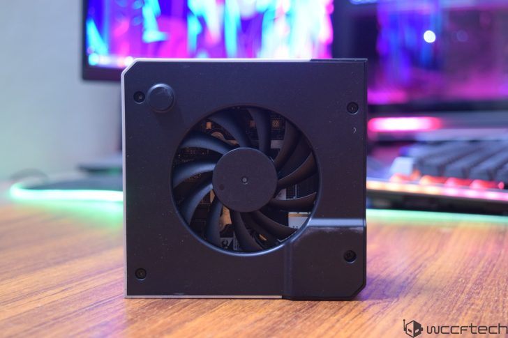 A compact, black unbranded fan-equipped computer component is placed on a wooden desk with a colorful LED-backlit background.