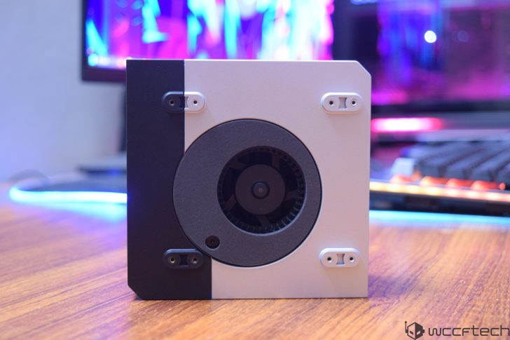 A close-up of a small, rectangular device with a black and white front panel and a central circular vent, placed on a wooden desk with a blurred background of a keyboard and monitor.