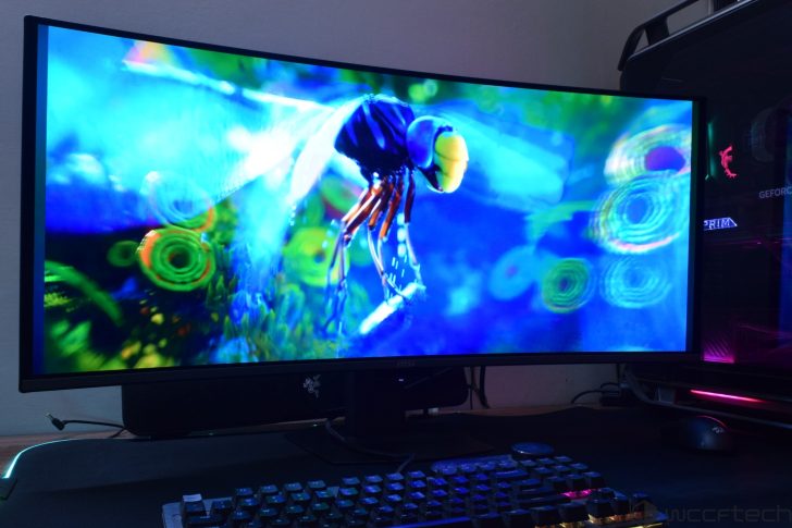 MSI MPG 341CQR X36 QD-OLED Gaming Monitor Review: Ultra-Wide Perfection With 360Hz A Razer logo is visible on a monitor displaying a vibrant, colorful digital artwork of an insect, with an MSI keyboard below and an illuminated PC housing with visible 'GEFORCE' text nearby.