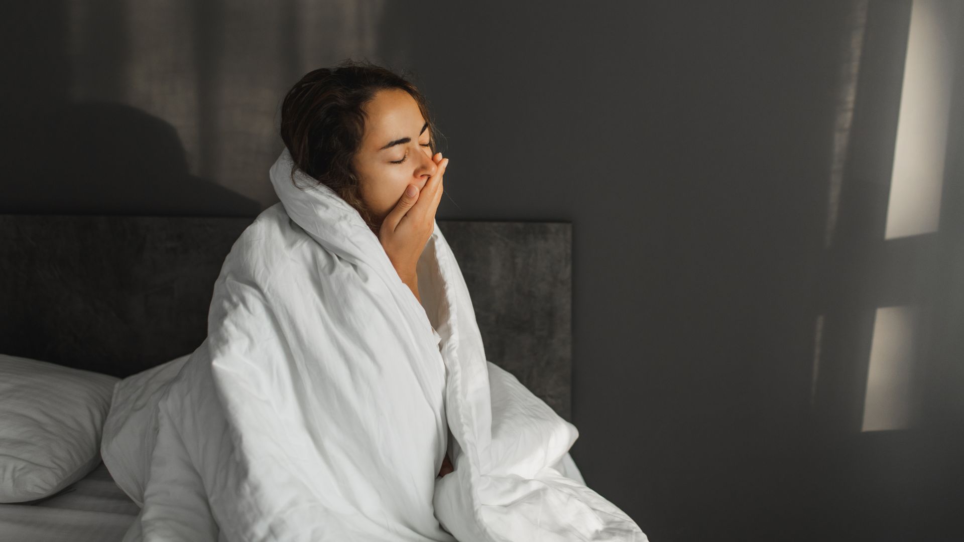 Μια γυναίκα κάθεται στο κρεβάτι καλύπτοντας το στόμα της με το χέρι της καθώς χασμουριέται. Έχει το λευκό της πάπλωμα τυλιγμένο στους ώμους της.