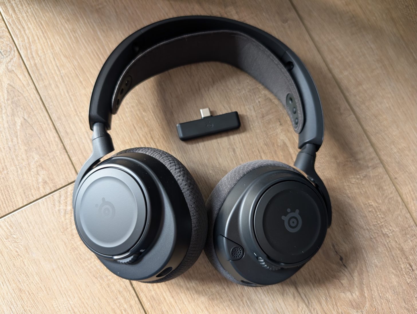 Ακουστικά SteelSeries με το λογότυπο SteelSeries στα ακουστικά, τοποθετημένα σε ξύλινη επιφάνεια δίπλα σε μια μικρή συσκευή USB.